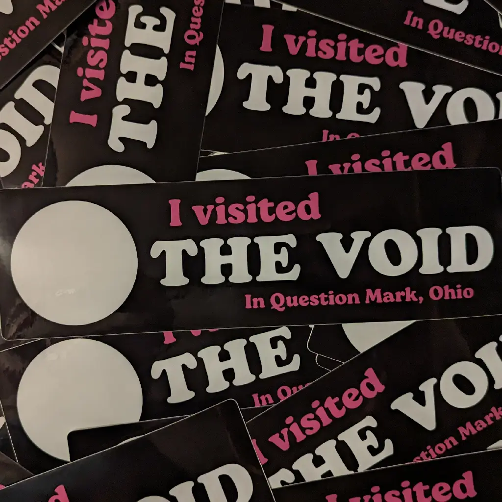 a pile of bumper stickers that read 'I visited The Void in Question Mark, Ohio' they are black bumper stickers, a large white circle is on the left side..